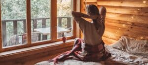 Woman stretching in a cozy wooden cabin bedroom with morning light streaming through a large window