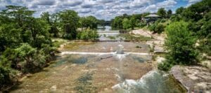 A scenic river with shallow waterfalls, surrounded by lush greenery and people relaxing on the bank