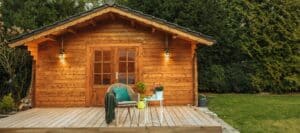 Small wooden cabin with a front porch, wicker chair, and potted plants surrounded by greenery