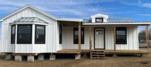 A newly constructed white modular house with a metal roof and a wooden porch stands on concrete blocks under a blue sky