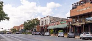 Bustling main street in San Marcos, Texas, featuring historic brick storefronts, parked cars, and signage for local businesses like Harper’s and The Porch