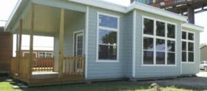 A small modular home with light blue siding and a wooden porch stands on concrete blocks, with large windows reflecting a bright sky