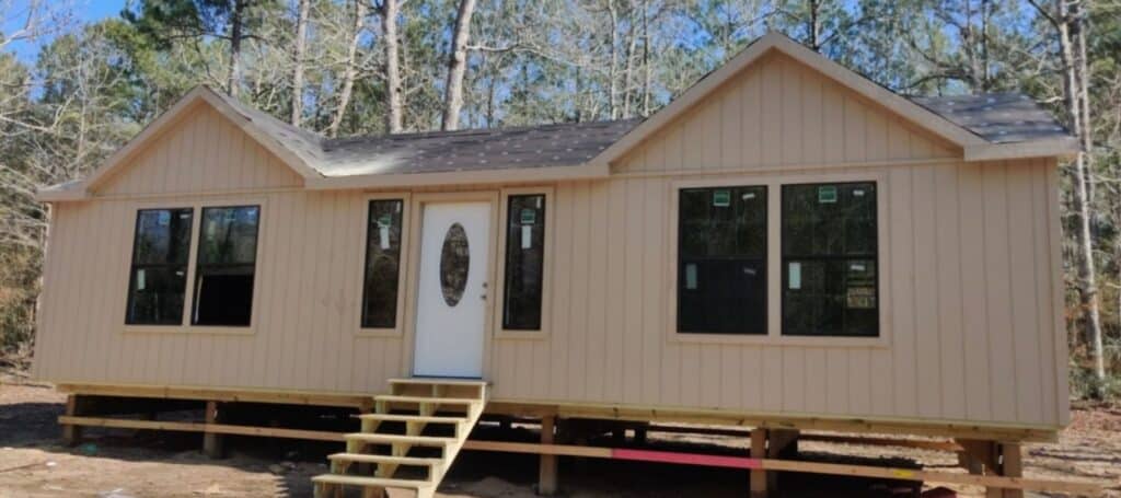 Light brown prefab cabin on raised wooden supports with front steps and black-trimmed windows
