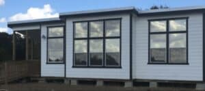 A modern modular home with white siding, black trim, and large windows sits on concrete blocks with a wooden porch and ramp