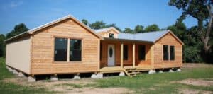 A newly built wooden modular house with a metal roof and a long front porch sits on concrete blocks in a field with green trees
