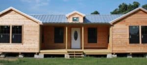 A newly constructed wooden modular house with a metal roof has a central porch with stairs and is supported by concrete blocks on a grassy lot
